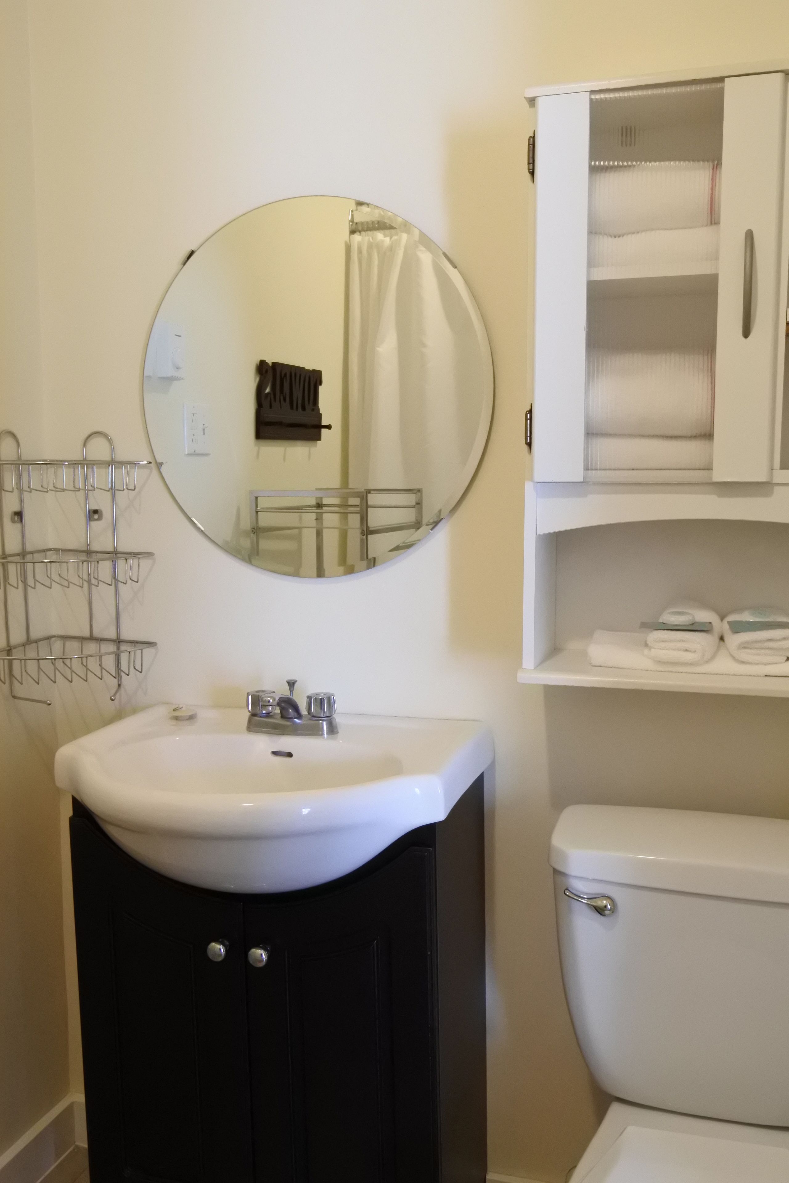 A bathroom with a white sink, a round mirror, a toilet, and a cabinet with towels.