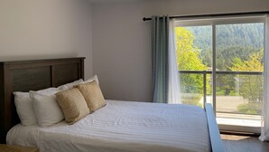 A bedroom with a large bed, a wooden headboard, and a view of a green landscape through a large window. - Black Rooster Guesthouse (Prince Rupert)