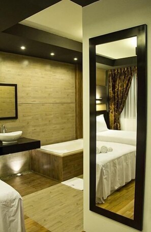 Een moderne hotelkamer met een houten vloer, een grote spiegel, een bed met wit beddengoed, een nachtkastje en een badkamer met een wastafel en een bad.