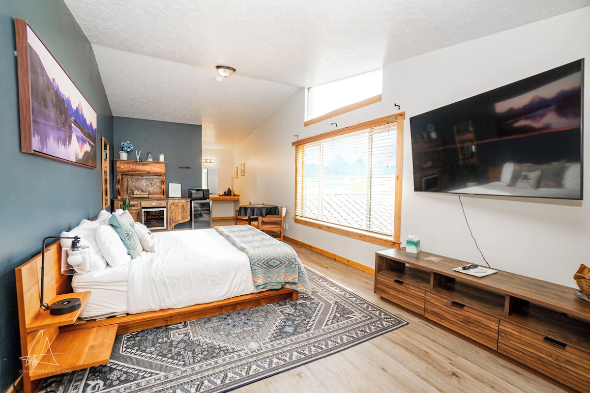 A bedroom with a large bed, a wooden dresser, a TV, and a window with blinds.