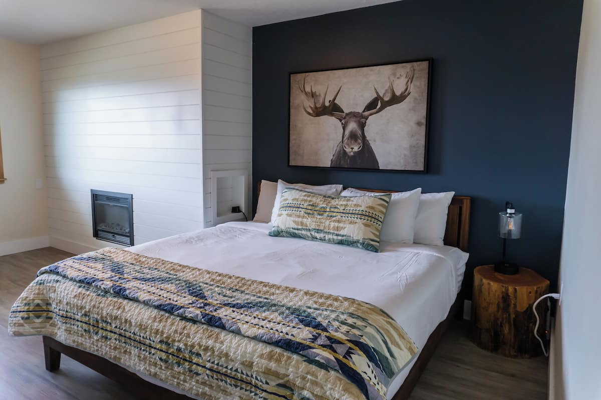 A bedroom with a large bed, a wall-mounted fireplace, a moose artwork, and a wooden nightstand with a lamp.