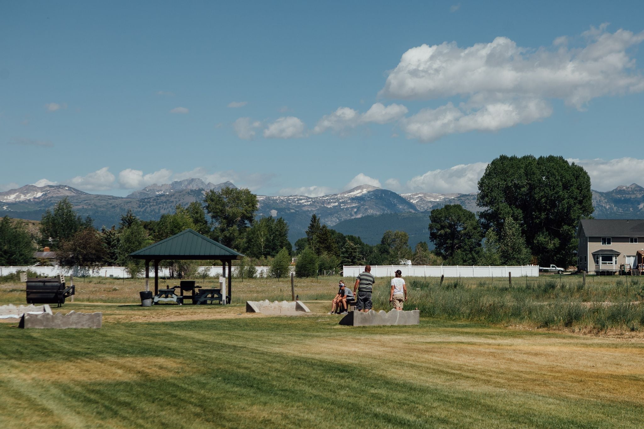 A grassy field with a gazebo, picnic tables, and a barbecue grill. In the background, there are mountains and a large tree.