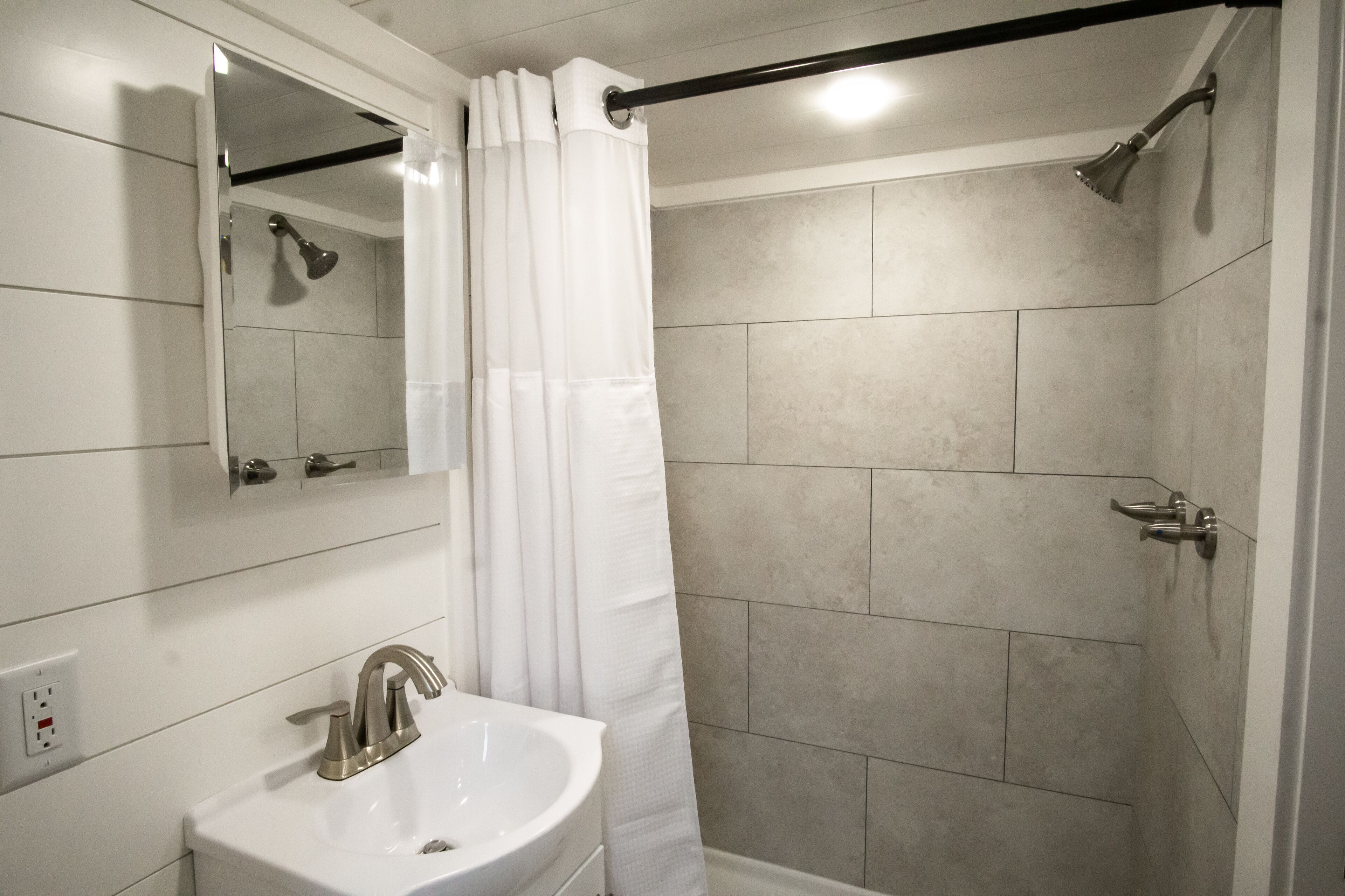Un baño con un lavamanos blanco, un espejo, una ducha con cortina y paredes de azulejos.