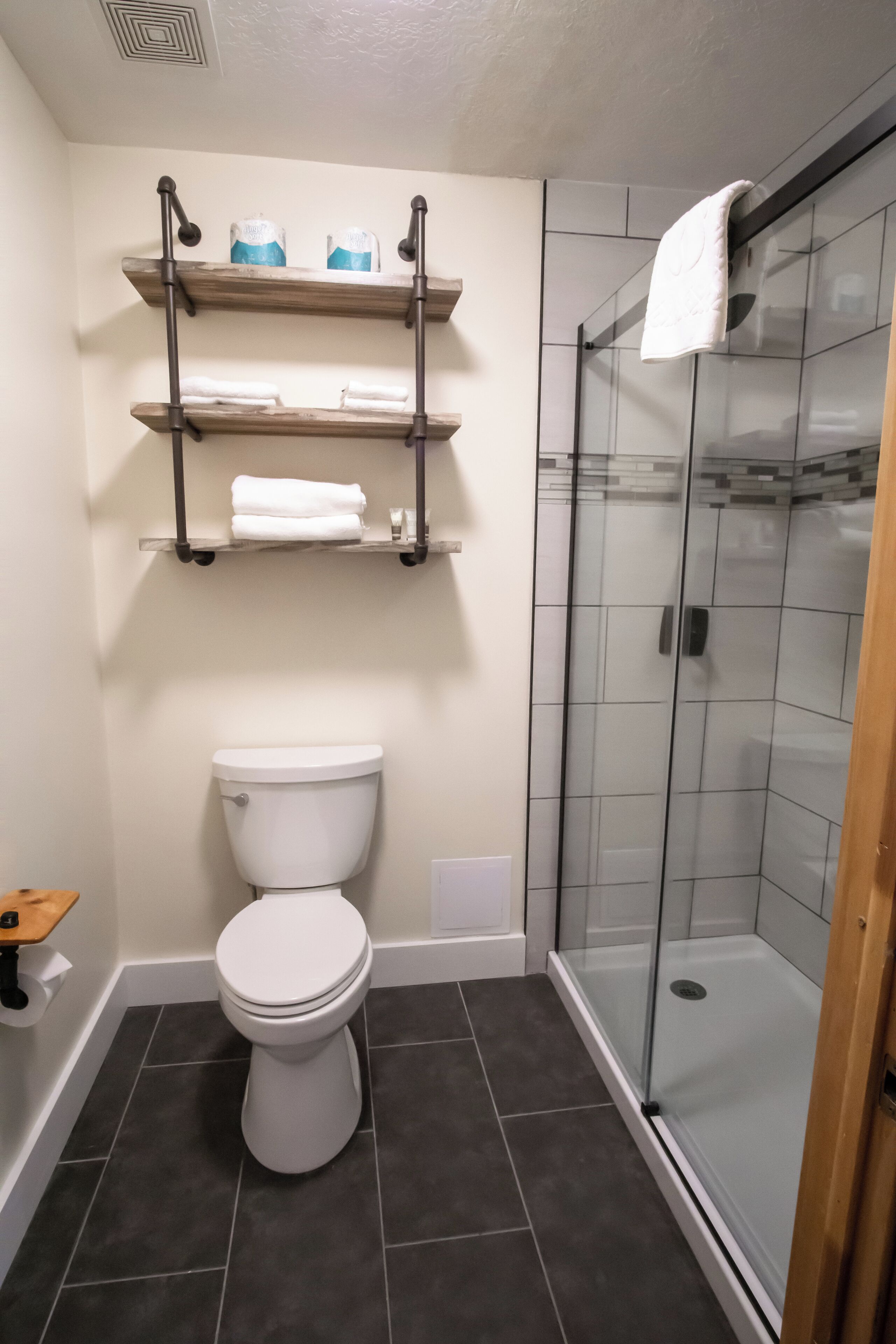 Ein Badezimmer mit Toilette, einer Duschkabine aus Glas und Holzregalen über der Toilette mit Handtüchern.
