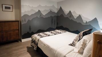 A bedroom with a mountain mural, a bed with patterned bedding, a wooden nightstand, and a ceiling fan.