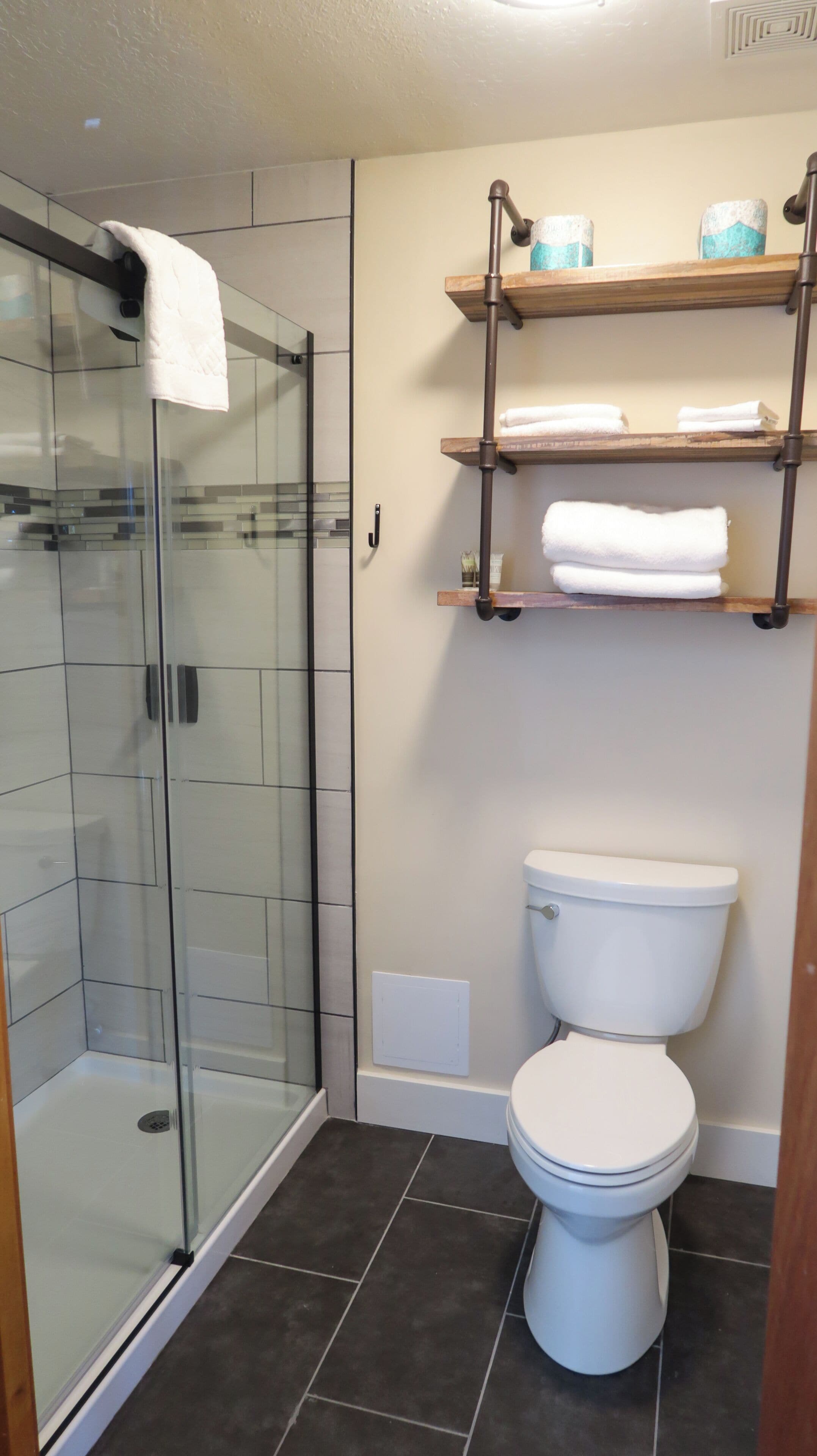 A bathroom with a glass shower enclosure, a toilet, and wooden shelves above the toilet.