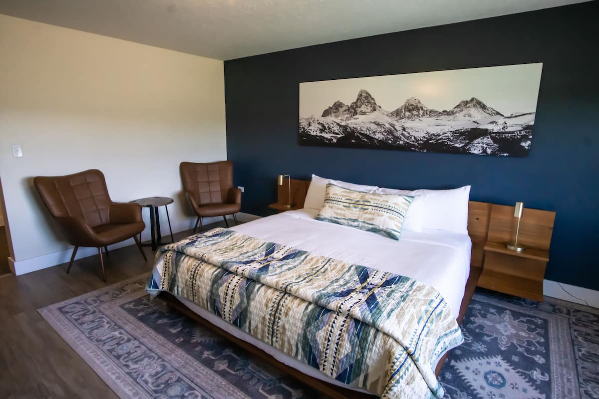 A bedroom with a large bed, two chairs, a small table, and a mountain landscape painting on the wall.