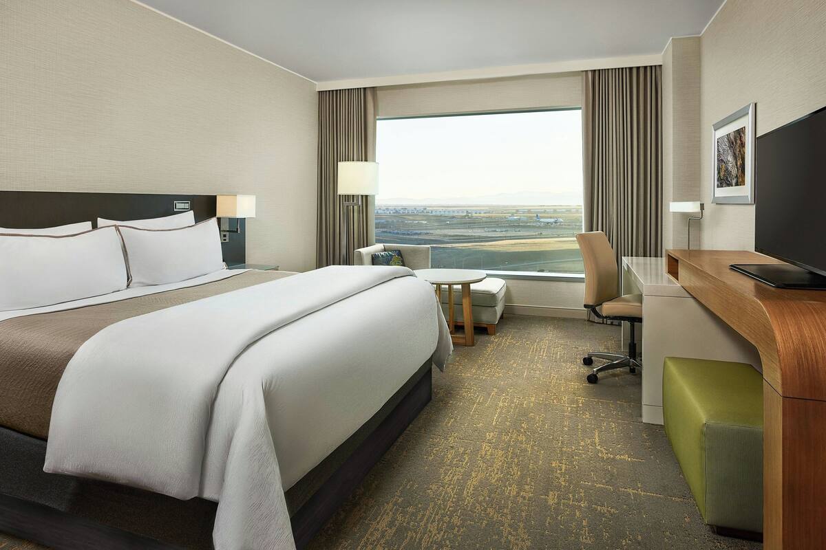 A hotel room with a large bed, a desk, a chair, and a view of the cityscape through the window.