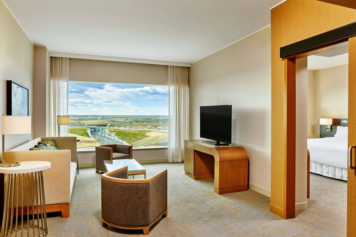 Hotelkamer met groot raam met uitzicht op een luchthaven en landingsbaan, een flatscreen televisie, een bank, een klein tafeltje en een bed.