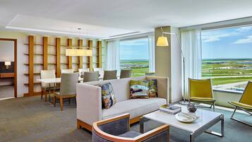 Habitación de hotel moderna con sofá, sillas, una mesa de comedor y vista al aeropuerto.
