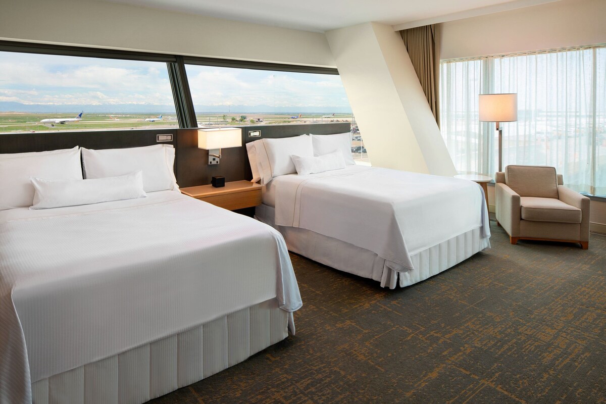 Hotelkamer met twee bedden, een nachtkastje, een lamp, een stoel en een groot raam met uitzicht op een luchthaven en het landschap.