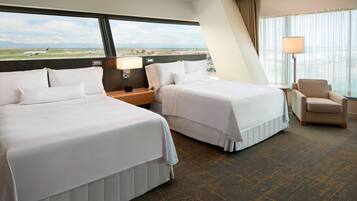 A hotel room with two beds, a nightstand, a lamp, a chair, and a large window offering a view of an airport and landscape.