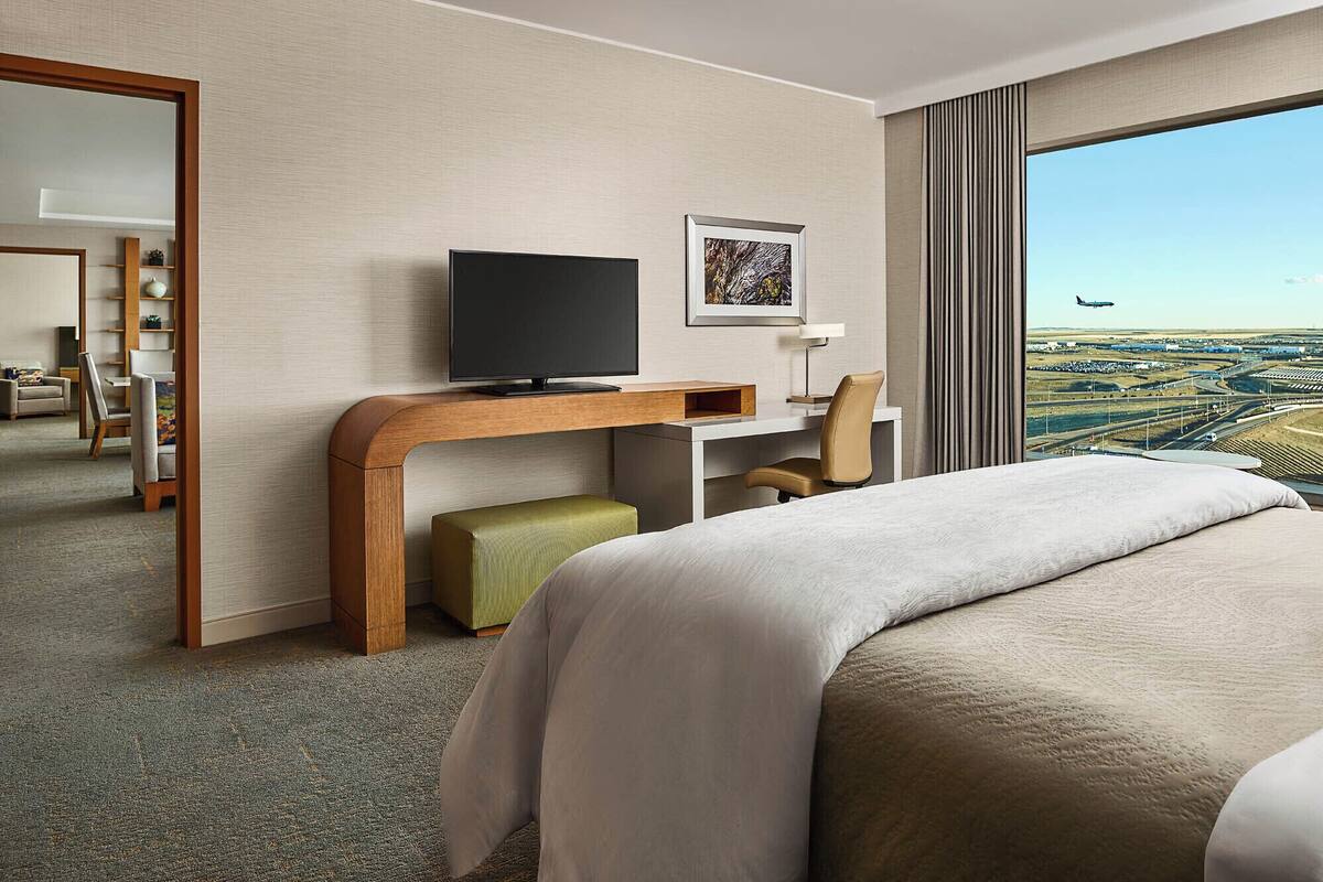 A hotel room with a bed, a desk with a TV, a chair, a small table, a mirror, and a large window with a view of an airport.