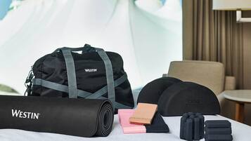 Una habitación de hotel con una cama, una bolsa Westin, una esterilla de yoga enrollada, una libreta rosa, una gorra negra y un juego de audífonos.