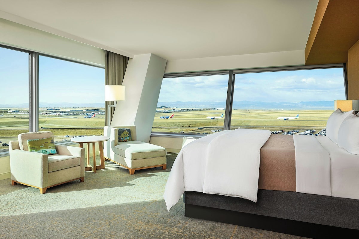 A hotel room with a large window offering a view of an airport and distant mountains.