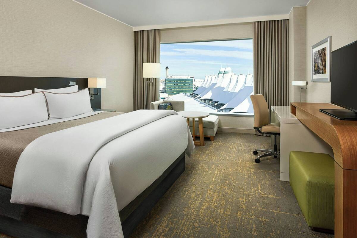 A hotel room with a large bed, a desk, a chair, a small table, and a view of the cityscape.