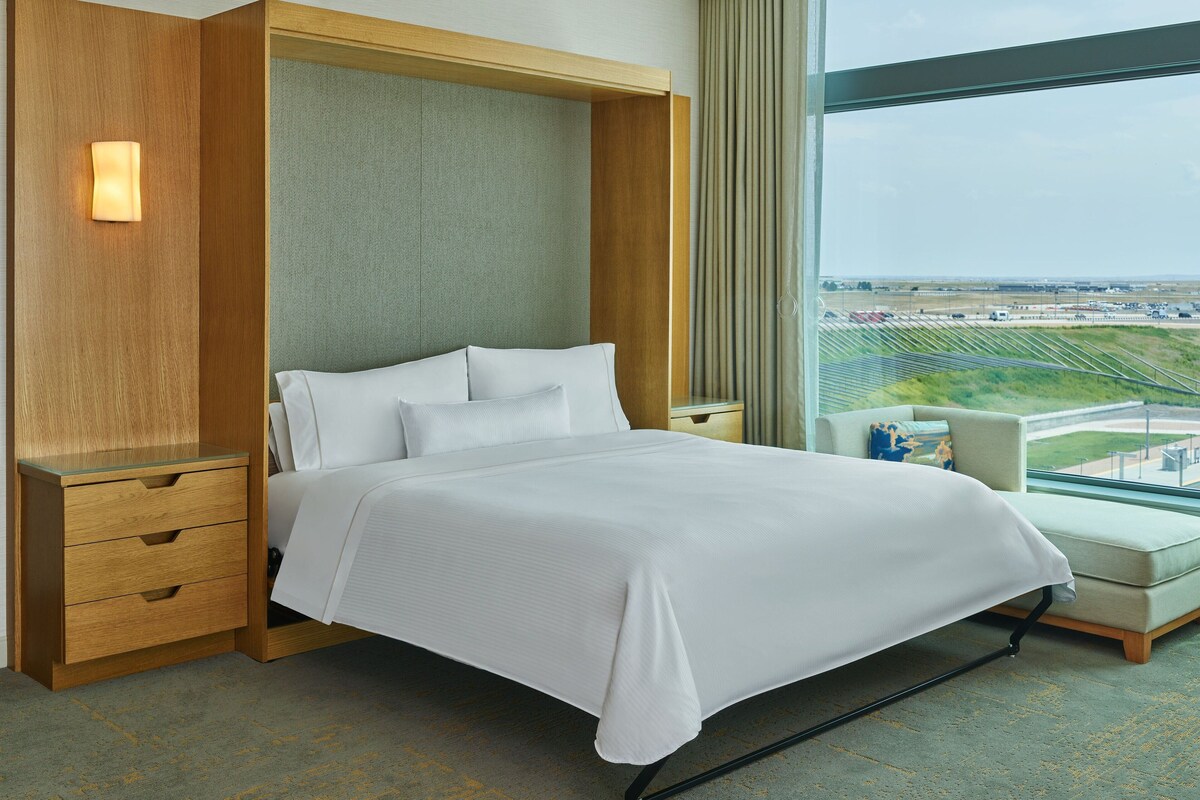 Hotelkamer met een groot bed, houten nachtkastjes en een zithoek met uitzicht op een vliegveld.