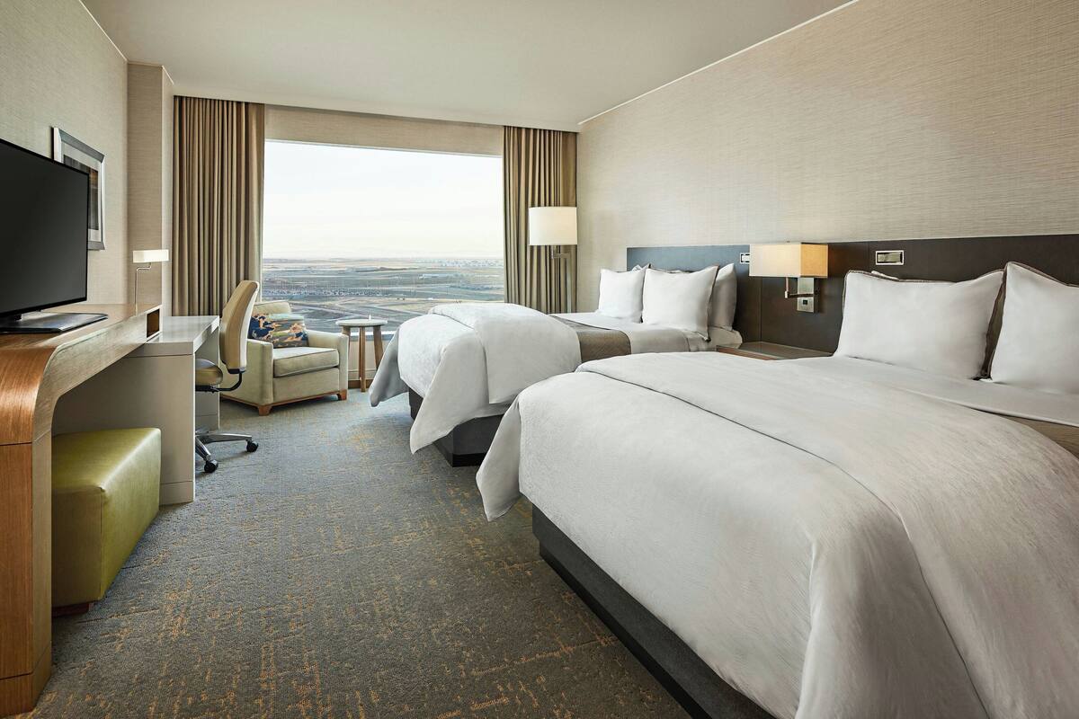 A hotel room with two beds, a desk, a chair, a TV, and a view of the city.