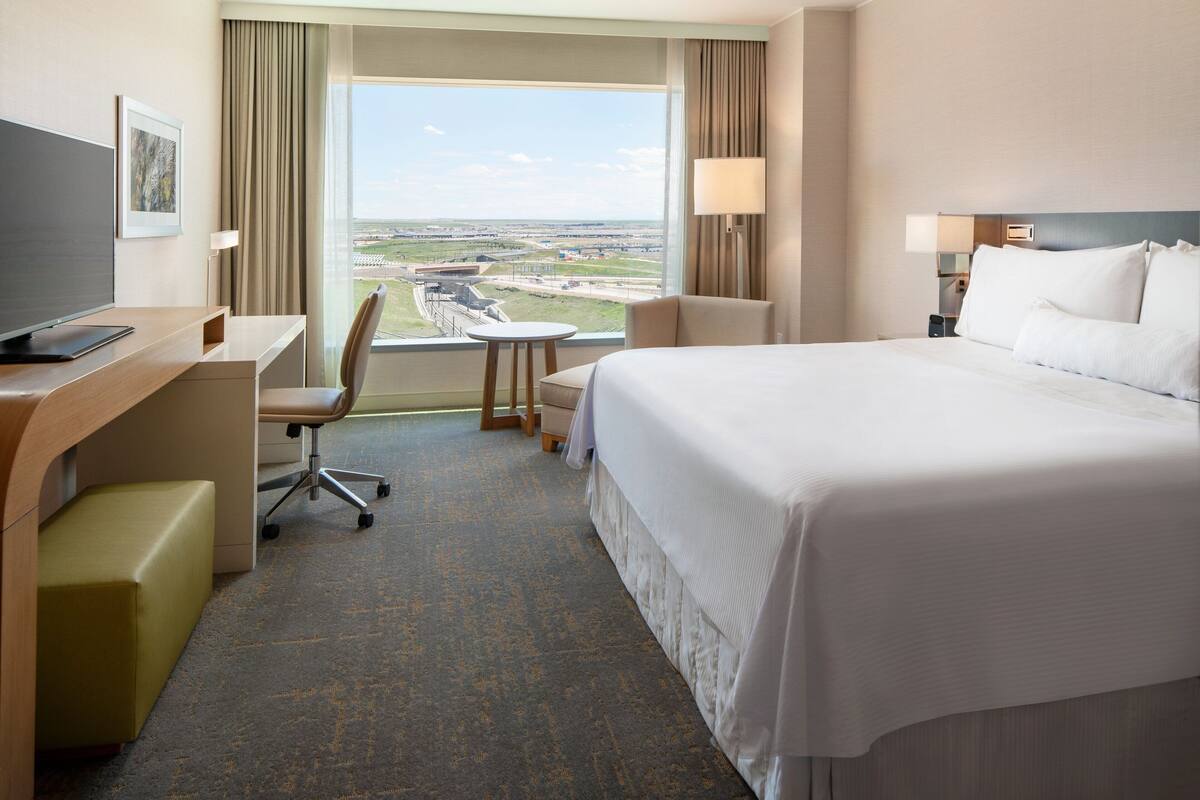 A hotel room with a large bed, a desk with a chair, a small table, and a view of the airport.