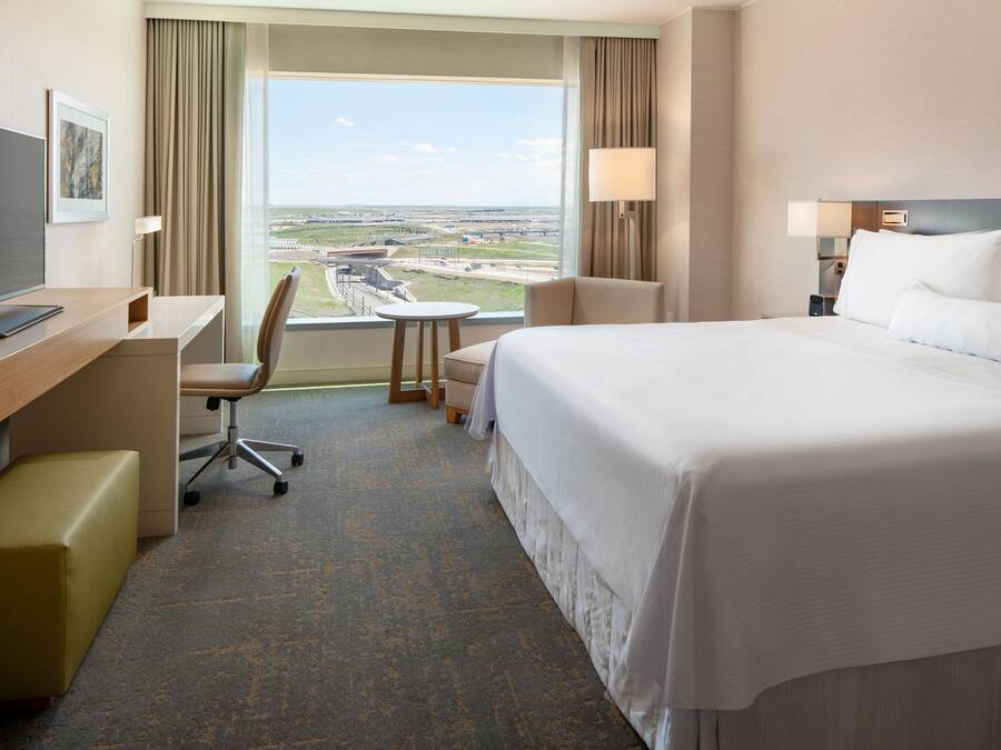 Ein Hotelzimmer mit einem großen Bett, einem Schreibtisch mit Stuhl, einem kleinen Tisch und Blick auf das Flughafengelände.