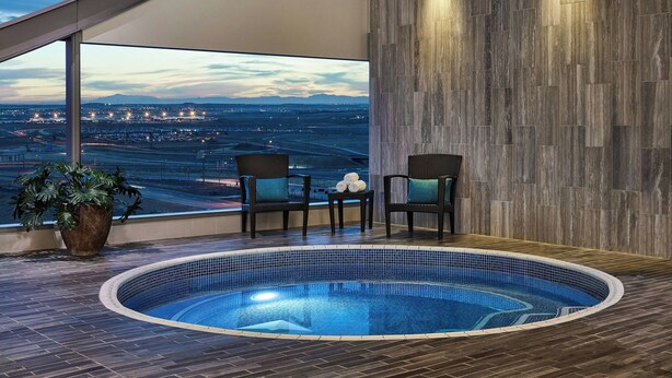 A modern indoor pool area with a view of a cityscape, wooden flooring, and two chairs with a potted plant.