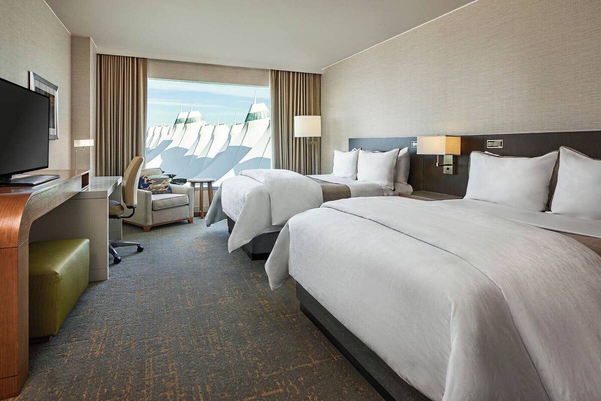 A hotel room with two beds, a desk, a chair, a TV, and a view of a modern building.