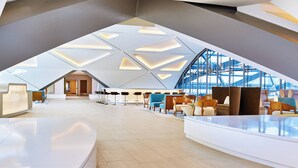 Een ruime lobby met een hoog plafond en geometrische lichtpatronen, modern zitmeubilair, en een groot raam met uitzicht op de omgeving.