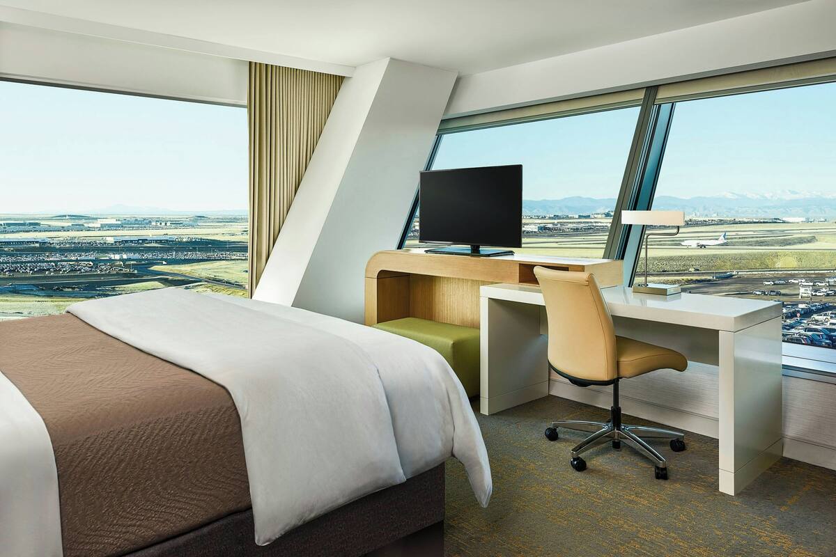 A hotel room with a large bed, a desk with a computer, and a view of an airport.
