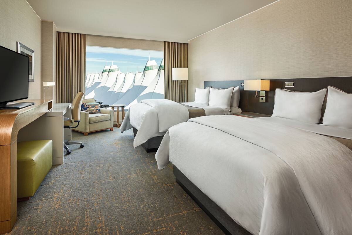 A hotel room with two beds, a desk, a television, and a view of sailboats.