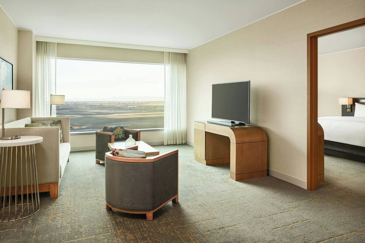 A hotel room with a large window, a bed, a desk with a TV, a sofa, and a small round table.