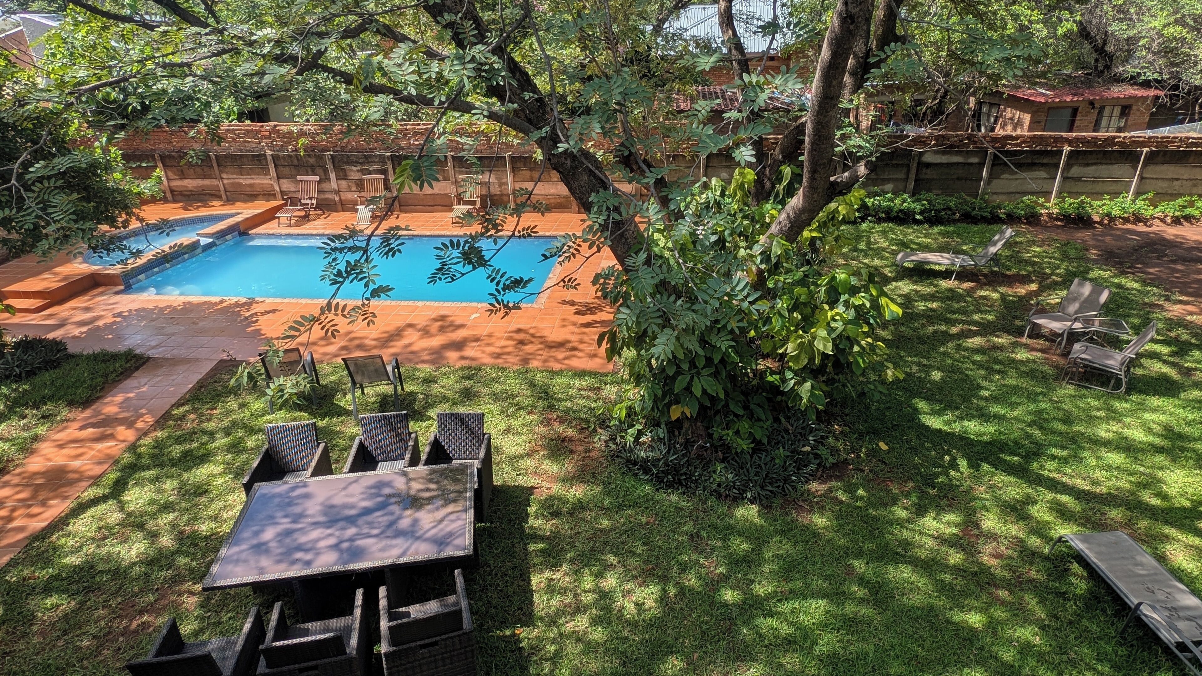 A backyard with a swimming pool, surrounded by a brick wall, and featuring a grassy lawn with a table and chairs.
