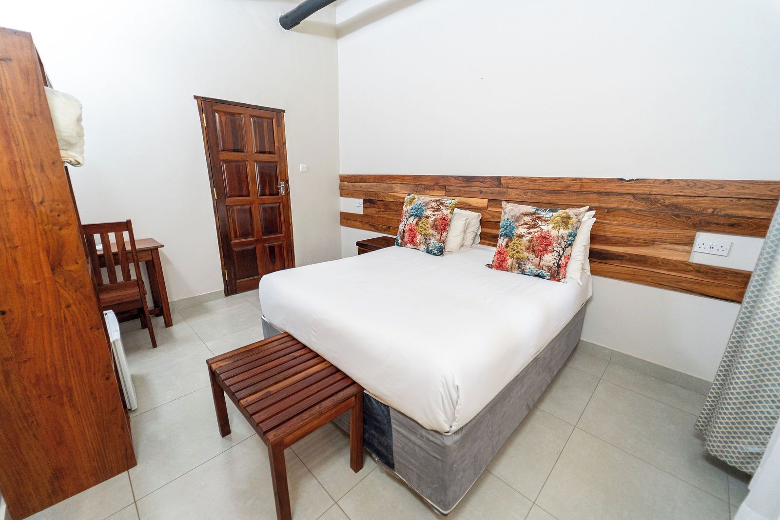 A bedroom with a wooden headboard, a bed with white bedding, two colorful pillows, a wooden bench, and a wooden door.