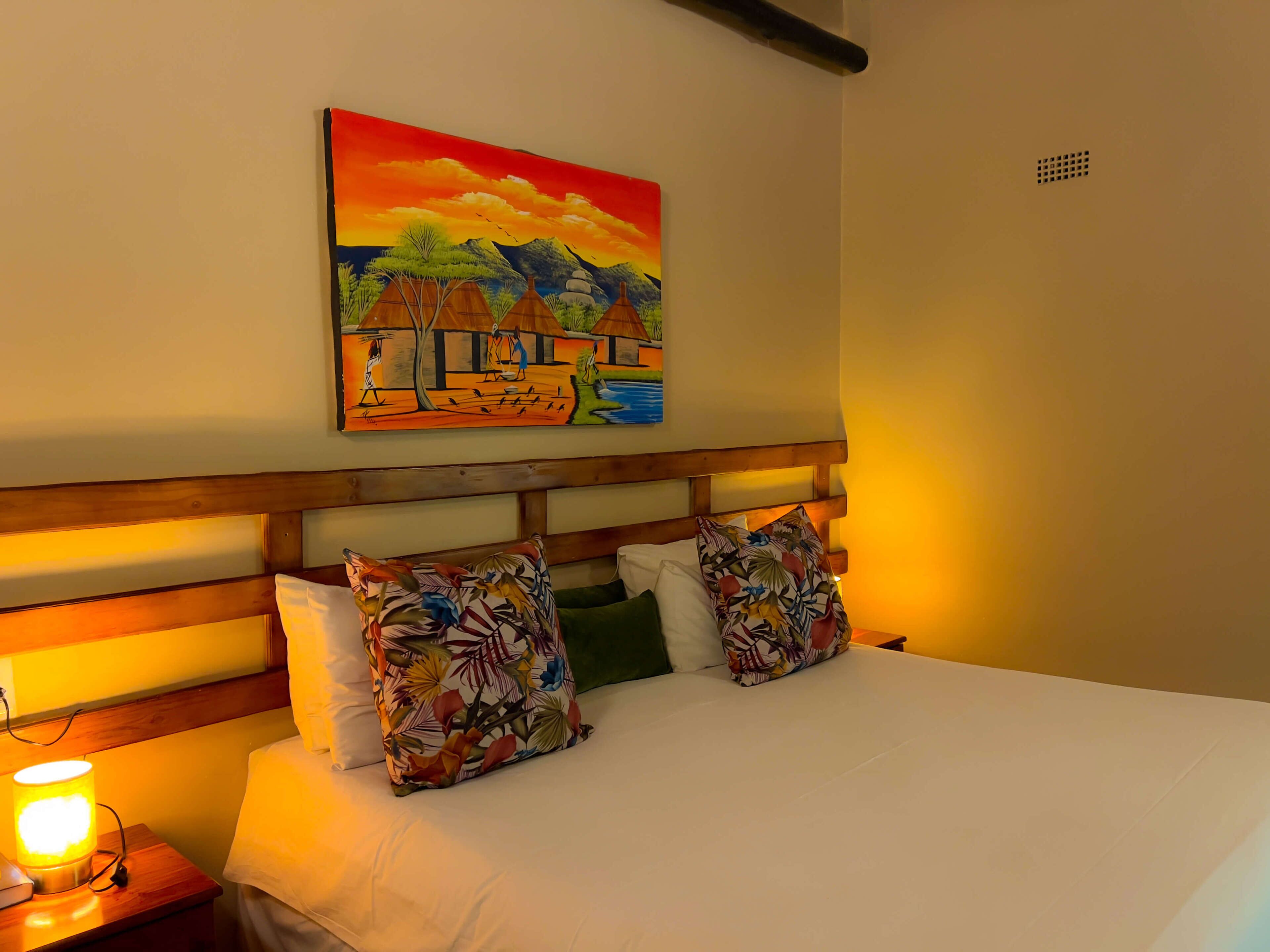 A bed with white linens, two colorful pillows, a wooden headboard, a nightstand with a lamp, and a painting of a landscape on the wall.