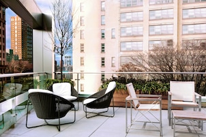 A balcony with wicker chairs and a table, overlooking a cityscape with tall buildings and a tree.