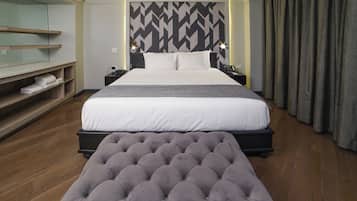 A modern hotel room with a large bed, a bedside table, a nightstand, a wall-mounted TV, and a seating area with a tufted ottoman.