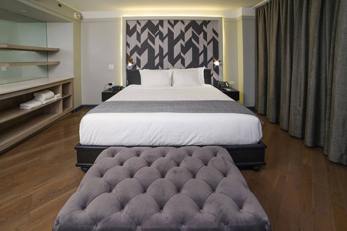 Una habitación de hotel con una cama grande, mesitas de noche, un televisor montado en la pared, un sofá y suelo de madera.