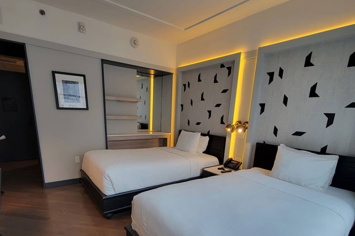 Habitación de hotel con dos camas, un espejo grande, una mesita de noche con lámpara y un cuadro enmarcado en la pared.