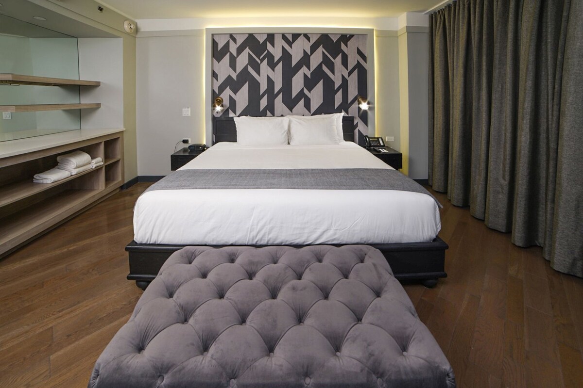 Una habitación de hotel con una cama grande, mesitas de noche, un televisor montado en la pared, un sofá y suelo de madera.