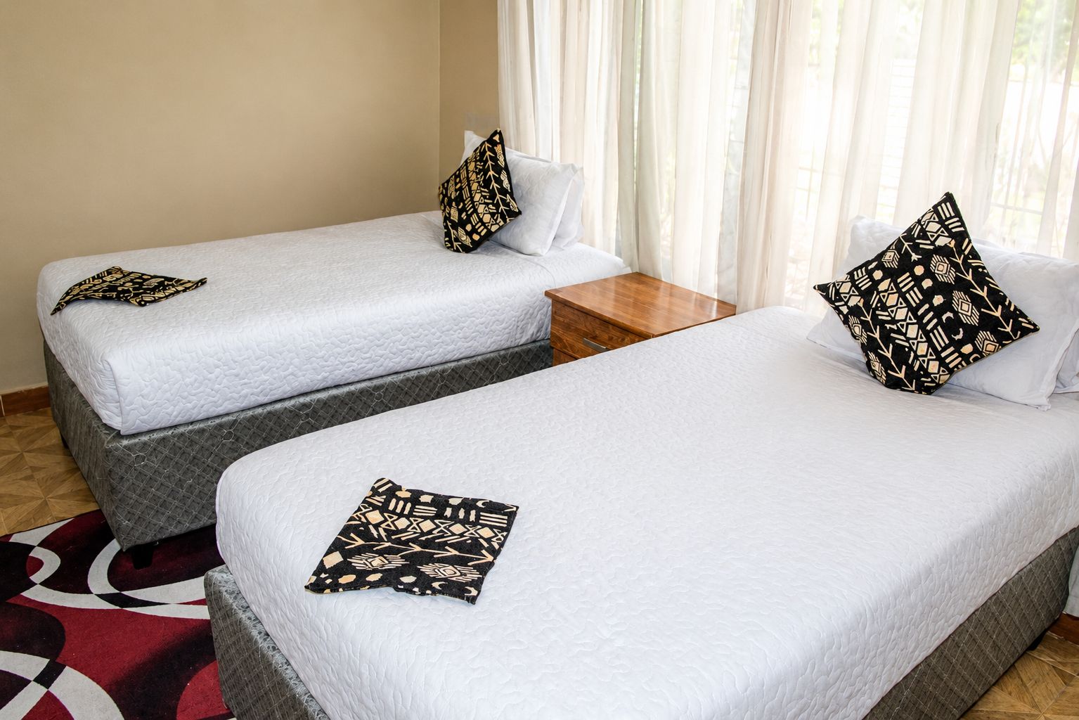 Two single beds with patterned pillows, a wooden nightstand, and a red and white patterned rug.