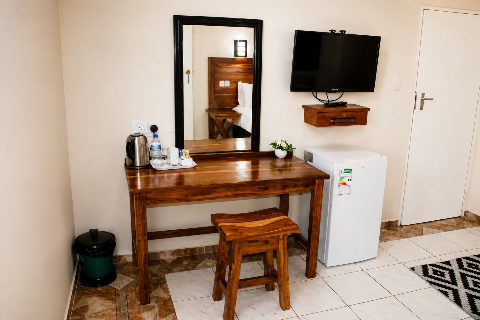 A compact hotel room with a wooden desk, a small stool, a television, a mini-fridge, and a trash bin.