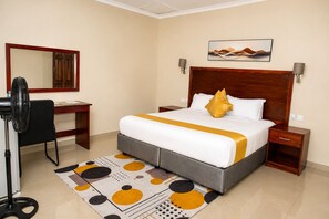 A hotel room with a bed, bedside tables, a desk, a chair, a fan, a mirror, and a wall-mounted artwork. - Wild Trekkers Lodge (Victoria Falls)