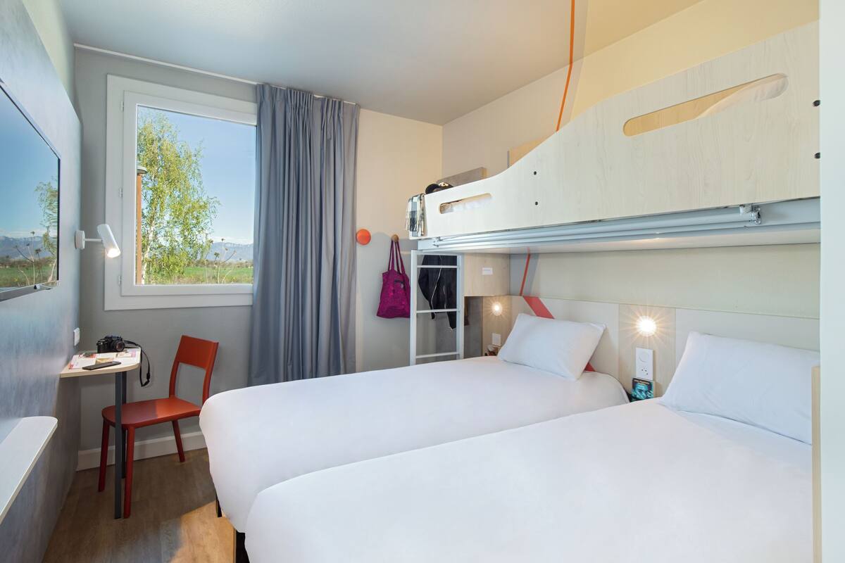 A hotel room with two beds, a desk, a chair, a window with curtains, and a view of the outdoors.