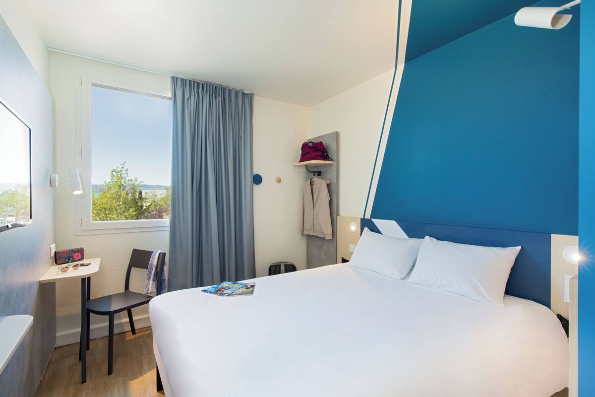 A hotel room with a bed, a desk, a chair, a window with curtains, a coat rack, and a view of the outdoors.