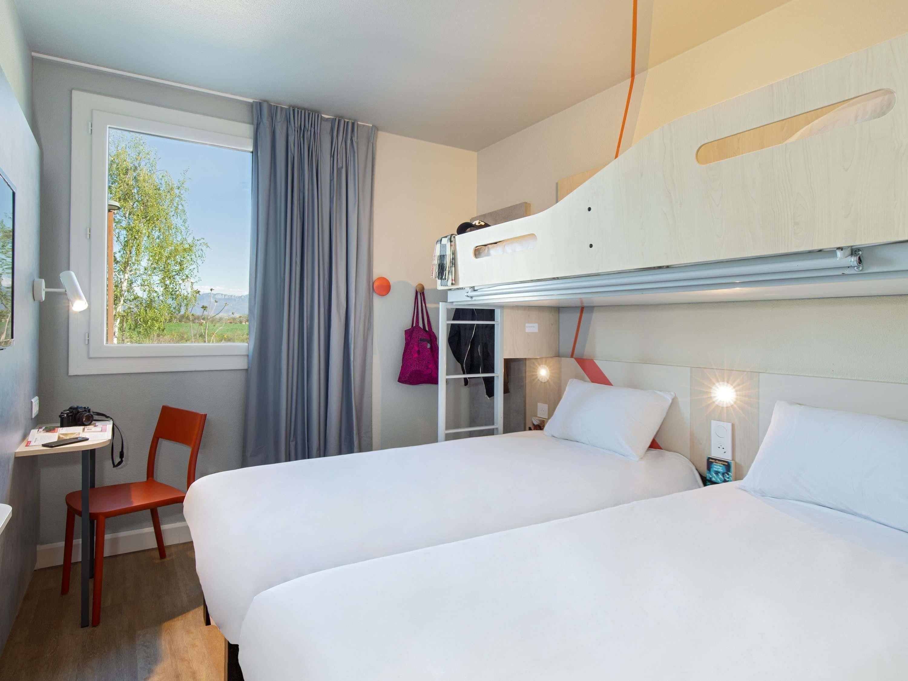 A hotel room with two beds, a desk, a chair, a window with curtains, and a view of greenery.