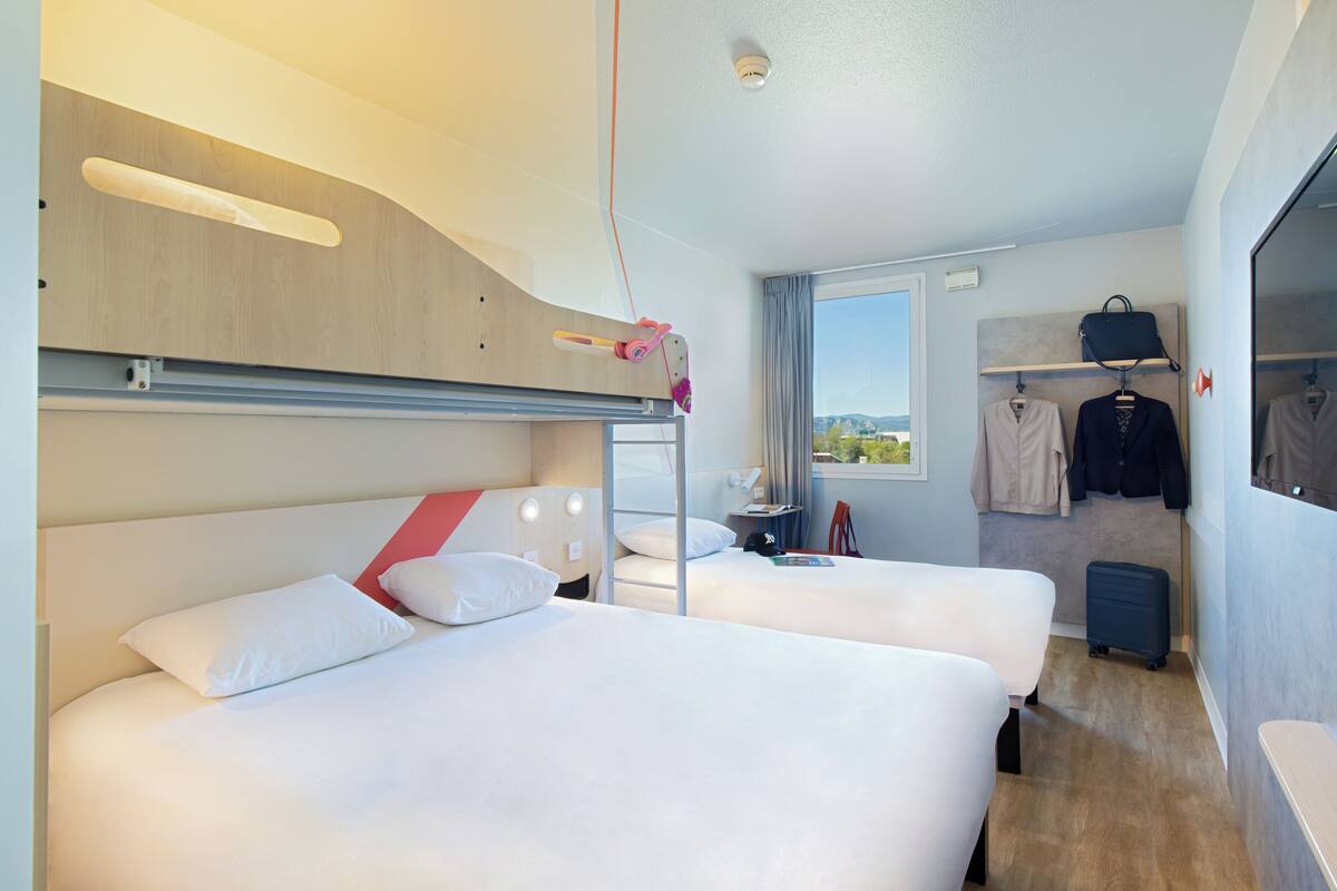 A hotel room with two beds, a desk, a window with a view, a coat rack, and a suitcase.