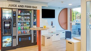 A juice and food bar with vending machines, a table, and seating areas. - ibis budget Valence Nord Plateau des Couleures (Saint-Marcel-les-Valence)