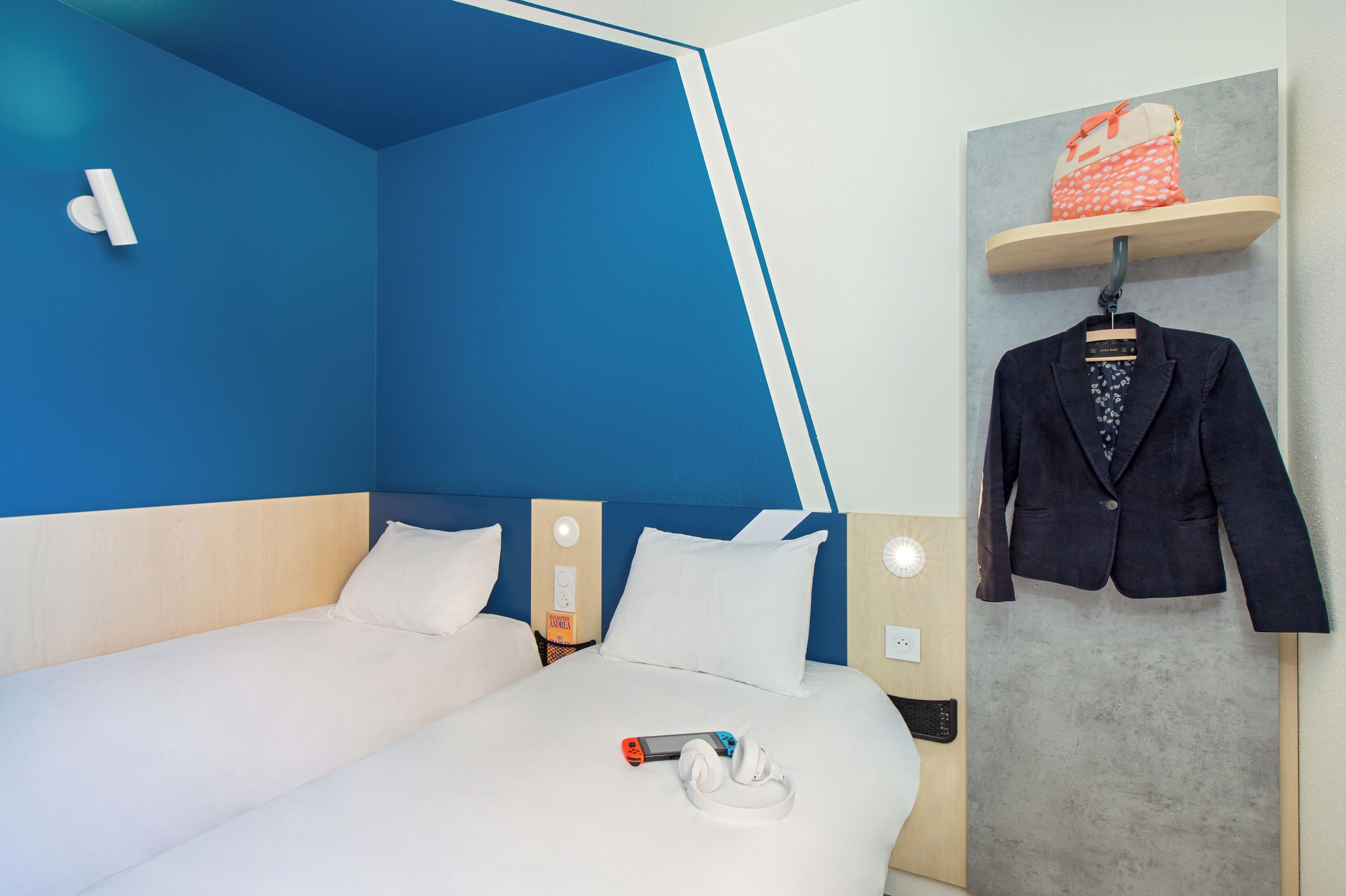 A hotel room with a bed, a nightstand, a wall-mounted shelf with a bag, and a hanging coat.