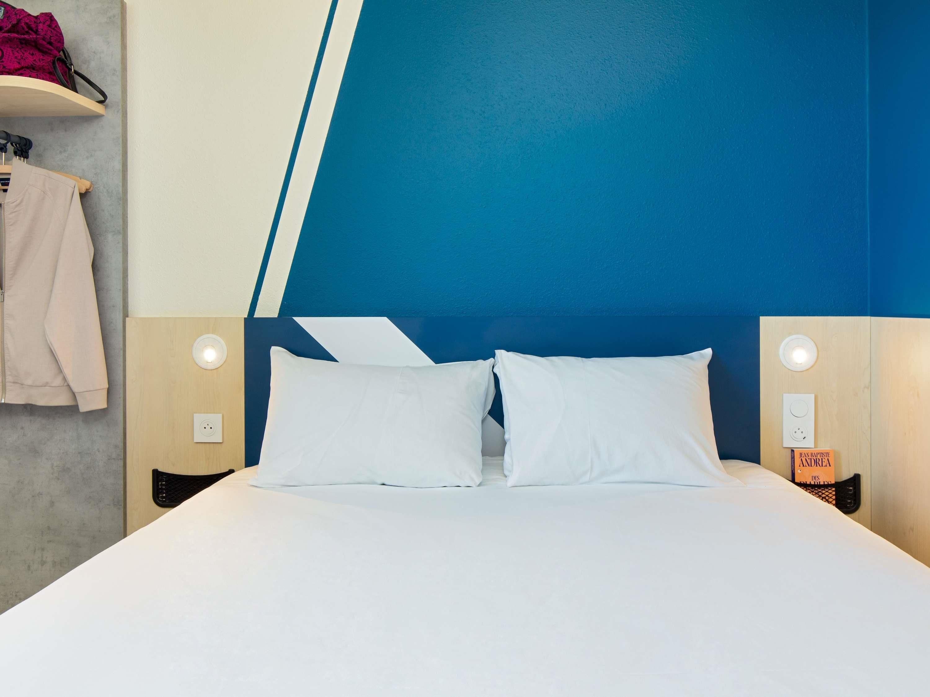 Habitación de hotel con cama, dos lámparas de noche, una mesita de noche y un estante en la pared con una chaqueta y un libro.
