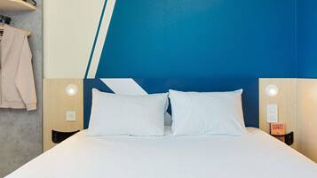 Habitación de hotel con cama, dos lámparas de noche, una mesita de noche y un estante en la pared con una chaqueta y un libro.