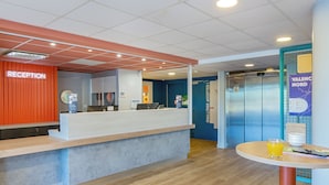 A reception area with a reception desk, a wall-mounted sign, an elevator, and a table with a glass of orange juice. - ibis budget Valence Nord Plateau des Couleures (Saint-Marcel-les-Valence)